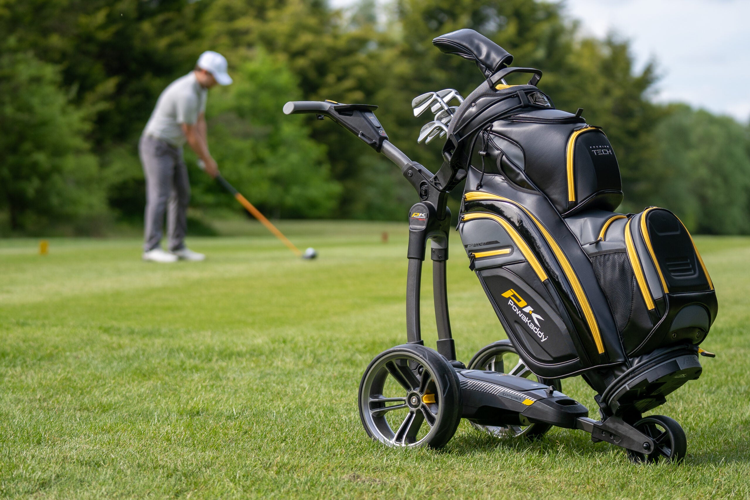 Golf E-Trolley mit Golftasche auf dem Fairway, Golfer schlägt Ball im Hintergrund